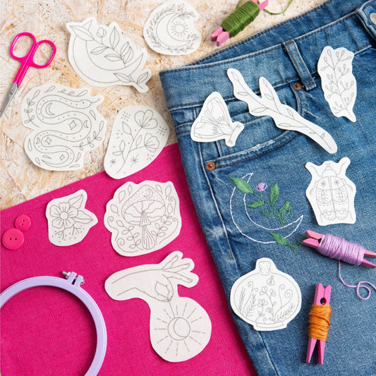 A styled flat-lay of embroidery or applique templates laid out on a bright pink surface next to blue denim jeans. Visible tools include pink scissors, thread spools, wooden buttons, a lavender embroidery hoop, and white paper templates with floral and abstract line designs.