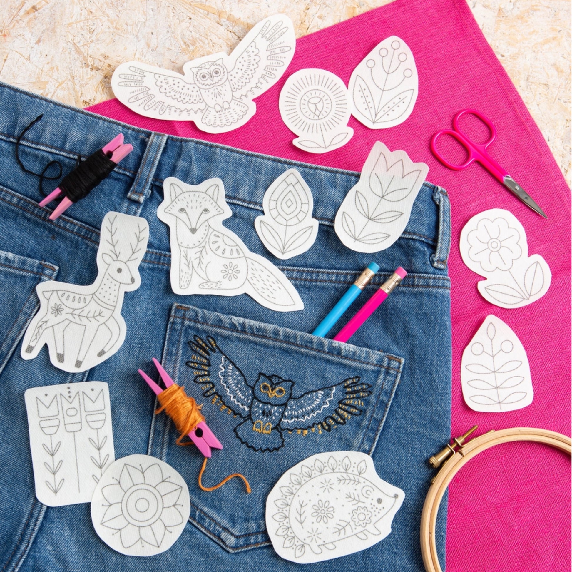A styled flat-lay of embroidered animal and floral design templates placed on blue denim jeans, with a bright pink fabric background, scissors, thread, pencils, and an embroidery hoop.