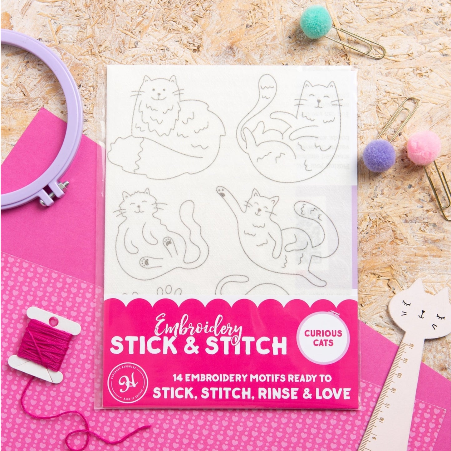 A package of 'Embroidery Stick & Stitch' featuring 'Curious Cats' embroidery motifs, displayed in a flat-lay arrangement with pink embroidery floss, an embroidery hoop, paper clips, and pom-poms on a wood background.