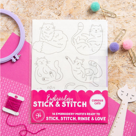 A package of 'Embroidery Stick & Stitch' featuring 'Curious Cats' embroidery motifs, displayed in a flat-lay arrangement with pink embroidery floss, an embroidery hoop, paper clips, and pom-poms on a wood background.