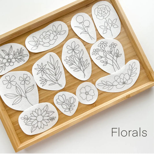A set of white fabric patches with delicate gray floral line drawings, arranged on a light bamboo tray. The word 'Florals' is printed in black text in the bottom right corner.