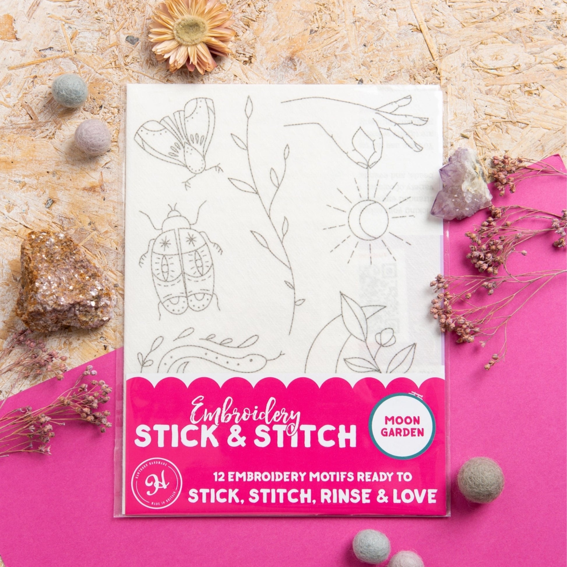 A packaged embroidery motif set labeled 'Moon Garden' and 'Embroidery Stick & Stitch' lies on a bright pink surface with decorative stones and dried flowers around it. The package shows a white sheet with gray outlined motifs including a butterfly, leafy stems, a sun, and a beetle.