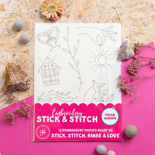 A packaged embroidery motif set labeled 'Moon Garden' and 'Embroidery Stick & Stitch' lies on a bright pink surface with decorative stones and dried flowers around it. The package shows a white sheet with gray outlined motifs including a butterfly, leafy stems, a sun, and a beetle.