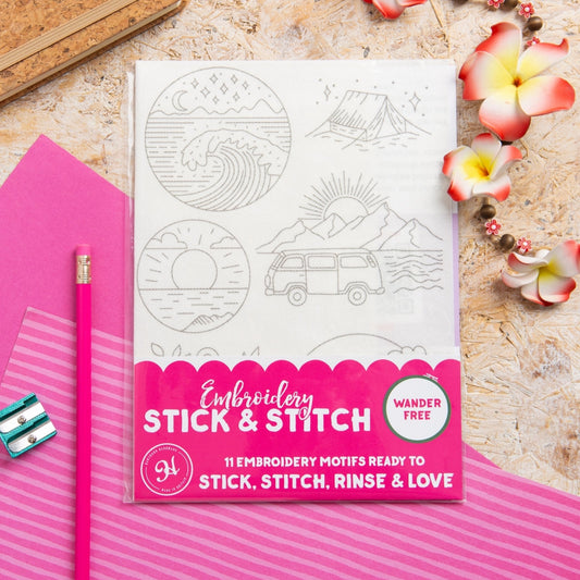 Embroidery Stick & Stitch kit featuring 11 embroidery motifs on a white sheet, styled in a flat-lay with a pink pencil, pink paper, and flowers on a textured beige background.