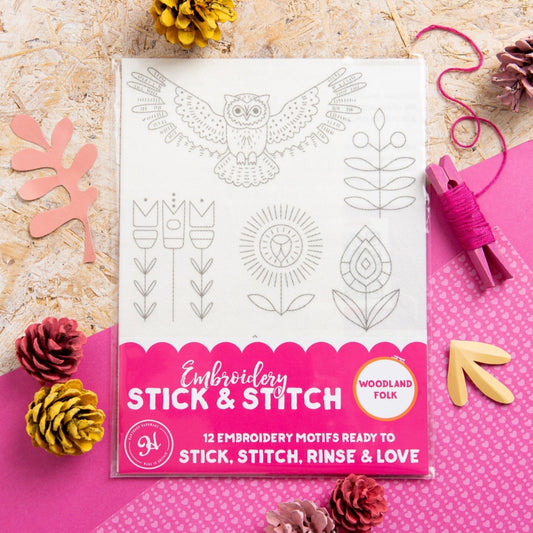 Embroidery Stick & Stitch kit in a clear plastic sleeve, featuring 12 woodland folk embroidery motifs on a white sheet with a bright pink bottom label. The package is styled in a flat-lay with pinecones, a pink clothes pin, and wooden leaf cutouts on a textured beige surface.