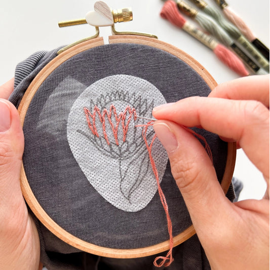 A close-up of hands holding a wooden embroidery hoop with dark gray fabric and stitching a coral-pink floral design. A few embroidery floss spools are visible in the blurred background.