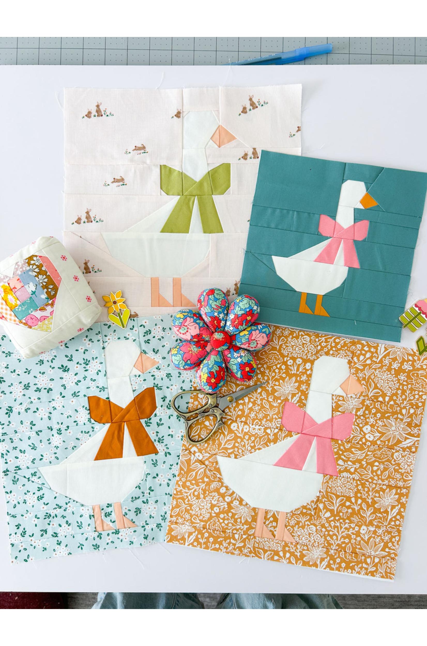 A collection of handmade quilted fabric blocks featuring a white goose design with different colored bows and scarves, arranged on a white surface alongside sewing scissors, a fabric pincushion, and a floral fabric bow.