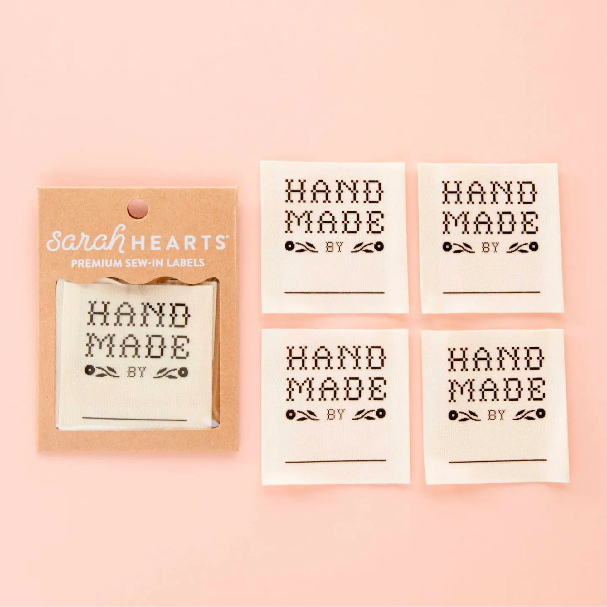 Four white fabric sew-in labels with black embroidered text reading 'HAND MADE BY' are displayed on a soft pink background, alongside a kraft paper packaging card branded 'Sarah Hearts'.