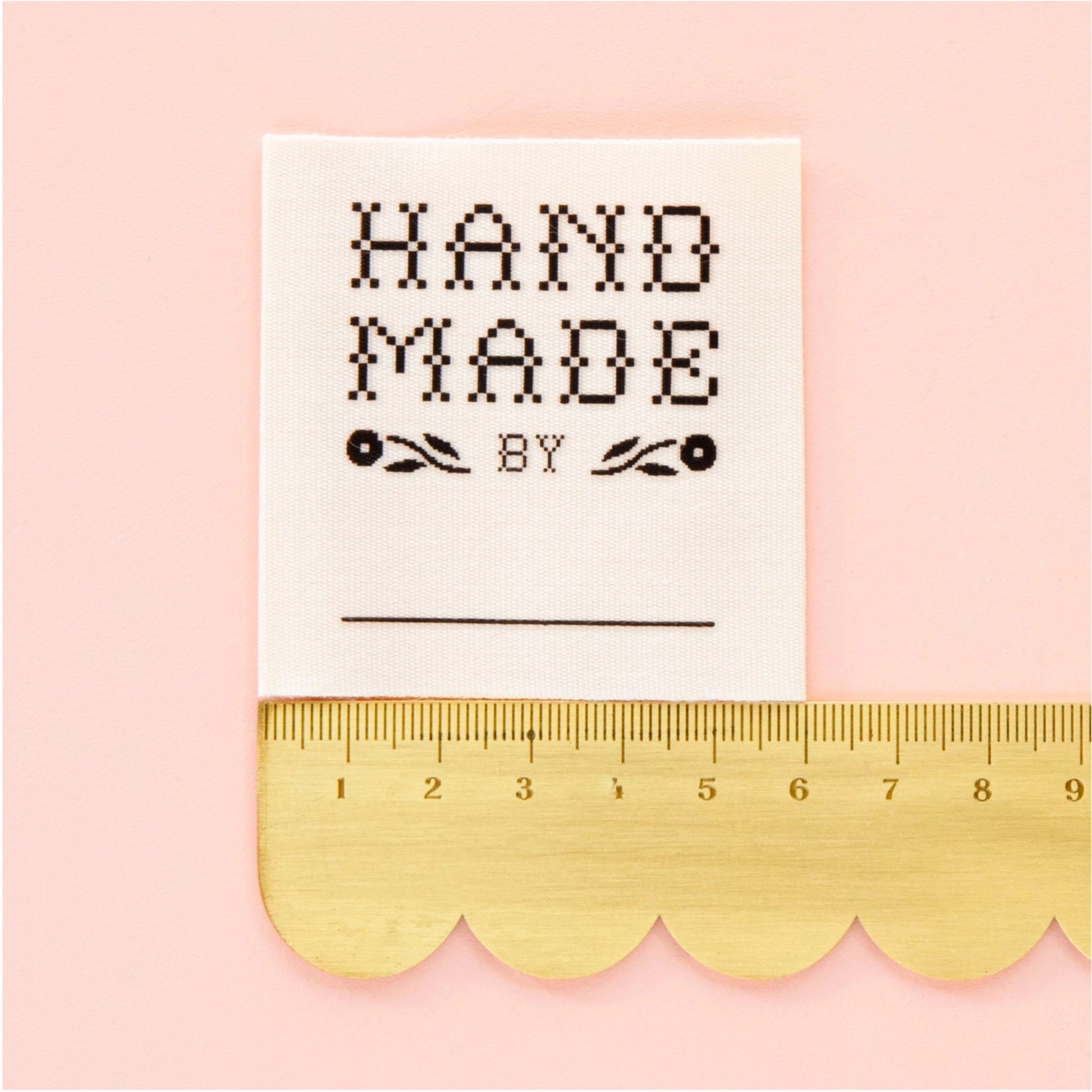 A square white fabric patch with black embroidered text reading 'HAND MADE BY' and a blank line below, shown next to a gold scalloped ruler on a light pink background.