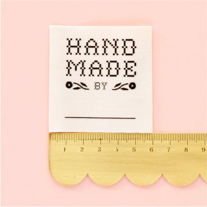 A square white fabric patch with black embroidered text reading 'HAND MADE BY' and a blank line below, shown next to a gold scalloped ruler on a light pink background.