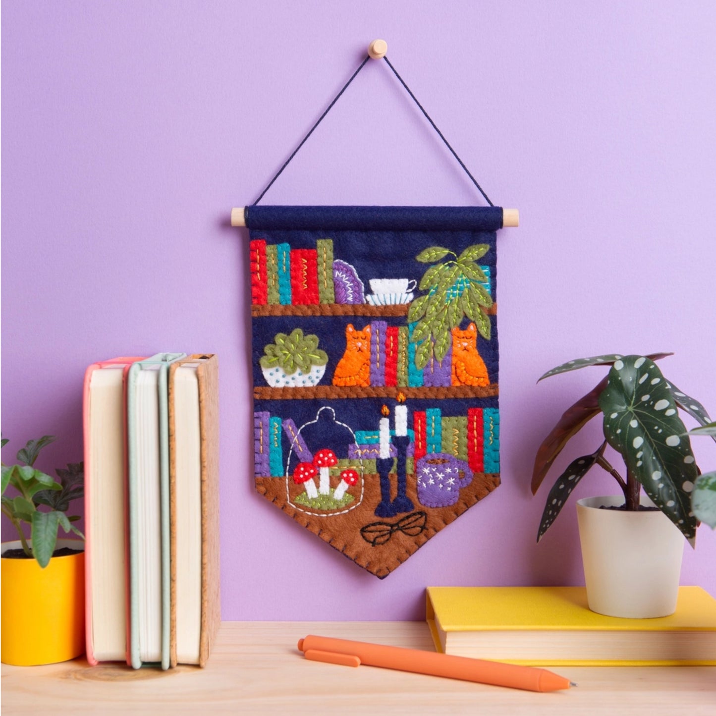 A colorful felt wall hanging with a bookshelf scene hangs on a lavender wall above a tabletop with books, a plant, and a pen.
