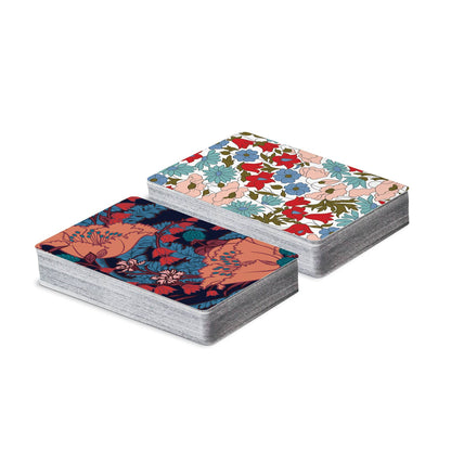 Two decks of patterned playing cards shown on a clean white background. One deck has a dark navy background with coral and teal floral motifs, and the other has a white background with red, blue, and peach floral motifs.